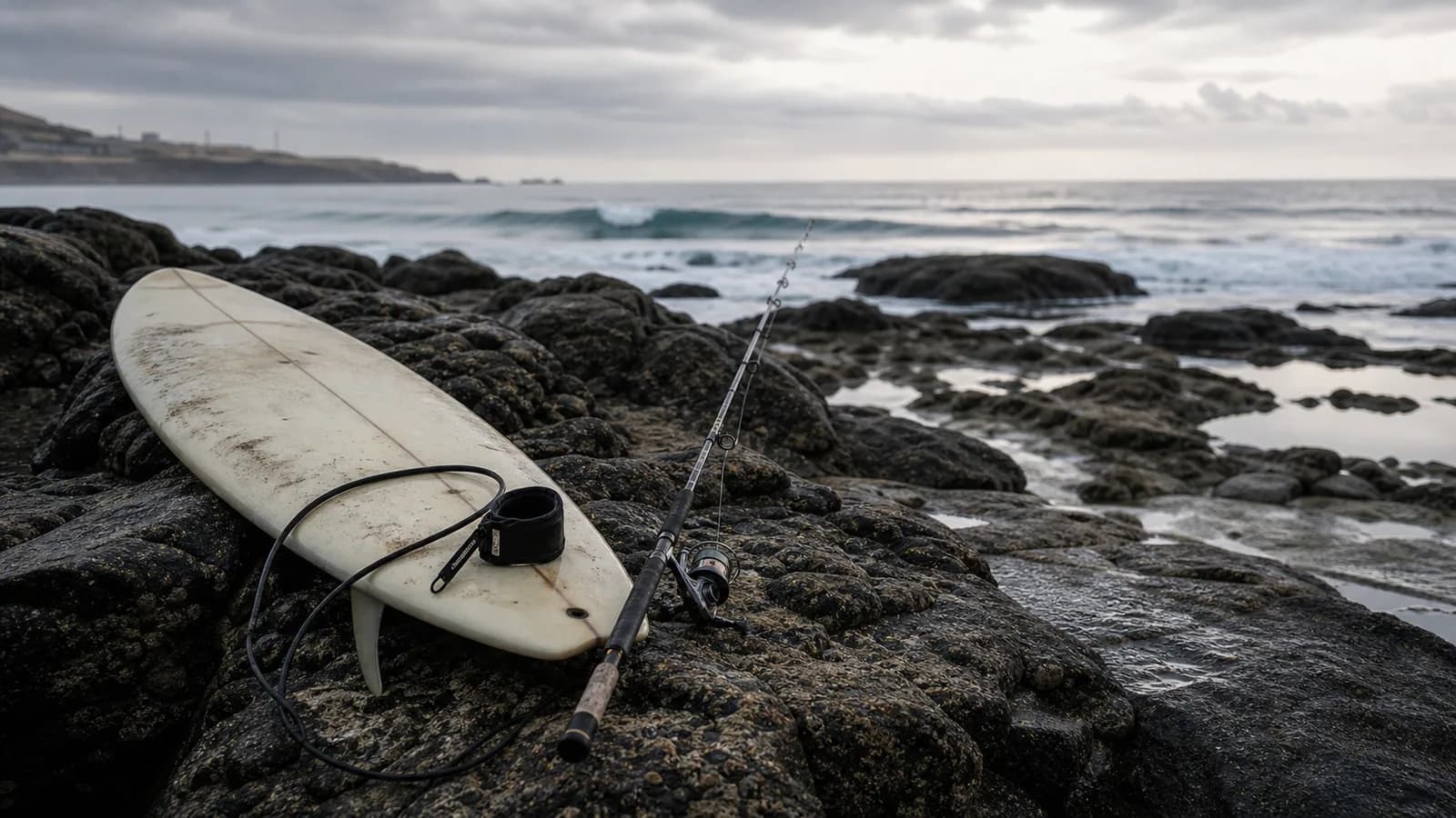 Salt-worn surfboard and fishing rod resting together beside Atlantic rocks